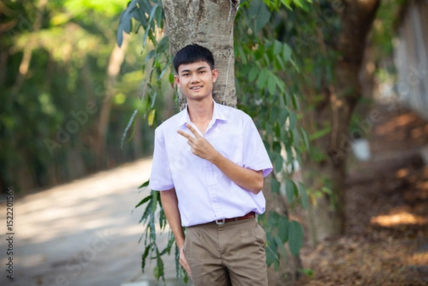 Fototapeta Thai boy student in official Thai school uniform (khaki or brown shorts), properly dressed, stand firmly in front of the tree with two fingers up, horizontal image