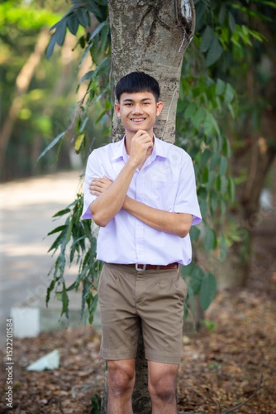 Fototapeta Thai boy student in official Thai school uniform (khaki or brown shorts), properly dressed, stand firmly in front of the tree with arms crossed, and one hand under his chin