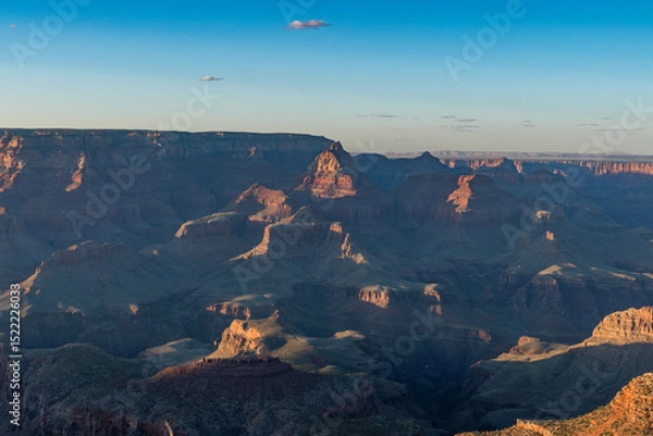 Obraz A breathtaking Grand Canyon sunset reveals layers of rugged red rock formations bathed in warm light and long shadows, while deep canyons and ridges form a mesmerizing view under a clear sky.