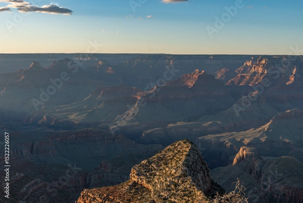 Obraz A breathtaking Grand Canyon sunset reveals layers of rugged red rock formations bathed in warm light and long shadows, while deep canyons and ridges form a mesmerizing view under a clear sky.