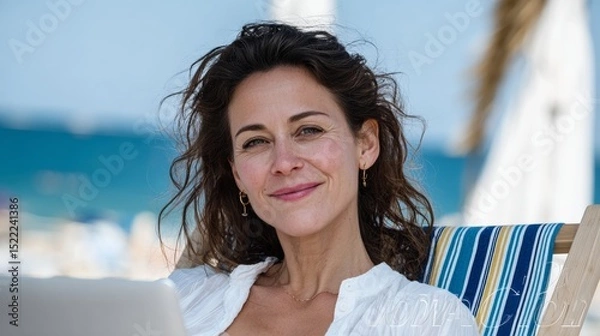 Fototapeta Woman is sitting on a beach chair with a laptop in front of her. She is smiling and she is enjoying her time