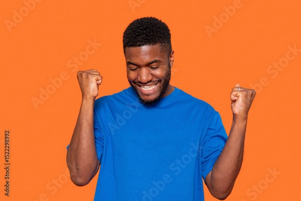 Obraz Joyful young man celebrates success with raised fists against a vibrant orange background, expressing happiness and excitement in a casual blue shirt