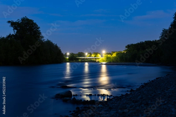Fototapeta A peaceful night scene of a calm river reflecting blue twilight and street lights from a distant bridge, surrounded by trees. Long exposure captures the serene ambiance of the evening