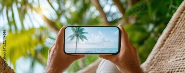 Fototapeta A person relaxing in a hammock views a tropical scene on their phone surrounded by greenery.