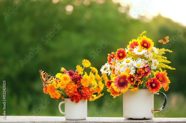 Fototapeta two enamel mugs with colorful flowers and butterflies on table in garden, natural background. summer season. rustic composition with flowers. relax time.