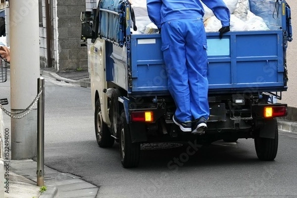 Fototapeta a Japanese garbage or utility truck, with a worker in blue overalls standing on the back, appearing to oversee the loaded materials or preparing for collection.A narrow urban street or alleyway,city l