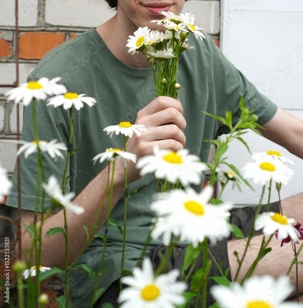 Obraz Farmer young man holding a bouquet of white garden daisies in his hands against the background of the wall of a stone house close-up as a concept of eco-friendliness