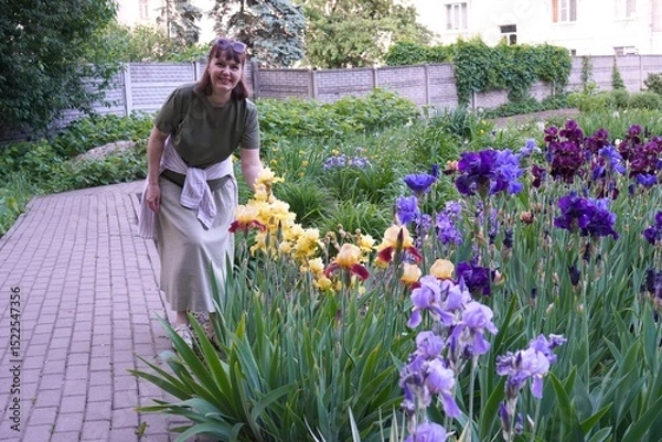 Obraz Happy woman florist standing near flower bed with colorful flowering irises in ornamental plant nursery in summer as botanical garden landscaping concept in summer