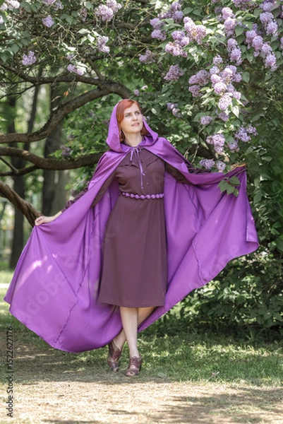 Fototapeta A beautiful young woman with red hair, against a background of blooming lilacs, in a cloak and hood. Medieval fantasy costume. Fairy tale heroine, princess, duchess.