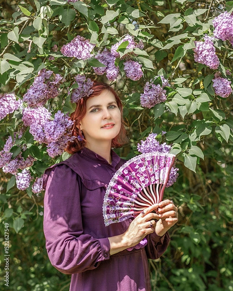Fototapeta A beautiful young woman with red hair, against a background of blooming lilacs, in a cloak and hood. Medieval fantasy costume. Fairy tale heroine, princess, duchess.
