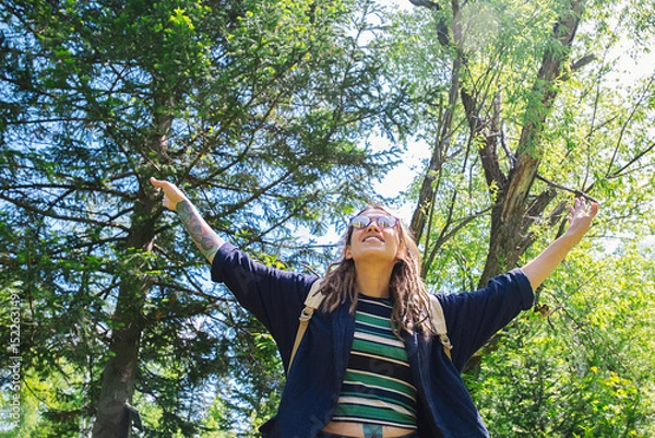 Fototapeta Woman Stands in a Forest, Her Arms Raised Up. Sunlight Filters Through the Trees, Creating a Vibrant, Natural Atmosphere.