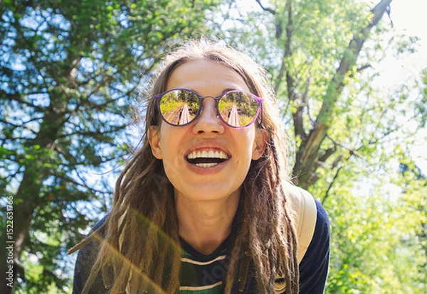 Fototapeta Smiling Woman with Long Dreadlocks and Sunglasses, Enjoying a Sunny Day in a Green Park Surrounded by Trees.