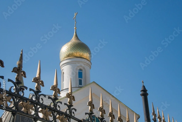 Fototapeta white brick Orthodox church with a golden dome and a cross against a clear blue sky, with a wrought-iron fence with pointed spires in the foreground