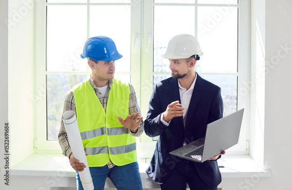 Fototapeta Portrait of young foreman discussing with professional architect design project or repair of new apartment on a laptop standing in empty room. Meeting with builder about interior decoration.