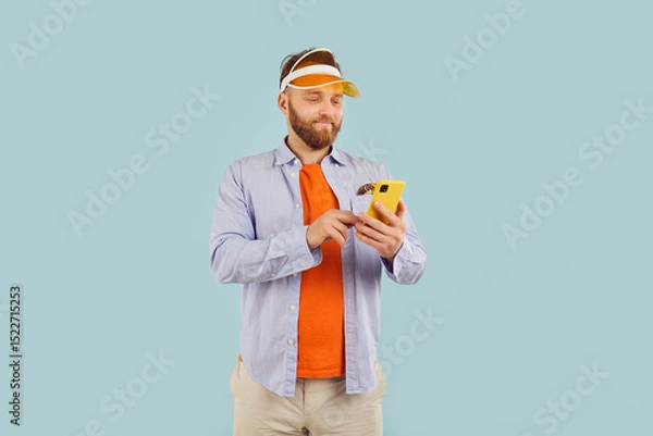 Fototapeta Connected and casual. Portrait of cheerful focused man in casual clothes engrossed in his smartphone. Smiling millennial man in sporting visor scrolls mobile phone screen on light blue background.