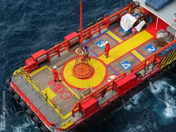 Obraz Workers on a vibrant red and yellow ship deck meticulously maneuver a large orange buoy using specialized equipment above the dark blue ocean waters.