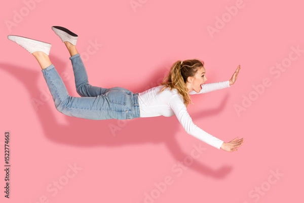 Fototapeta Young woman in casual attire appearing mid-air, striking a joyful and energetic pose against a vibrant pink background