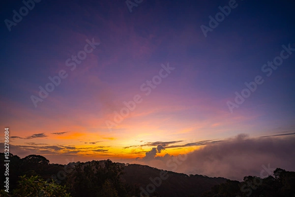 Obraz stunning mountainous landscape at sunrise with vibrant orange and pink hues illuminating the clouds against a blue sky. layer of mist settled in the valley, giving the scene a dreamy.