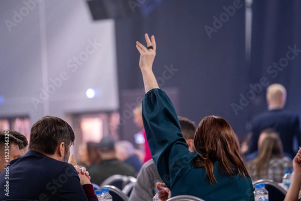 Obraz Person in crowded event raising hand to signal or ask a question. Back shot highlighting participation and engagement in a formal meeting or conference session.