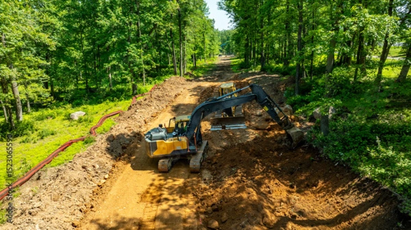Fototapeta Excavators work on clearing land in a densely wooded area. The operators maneuver heavy equipment to prepare the site for future development under clear blue skies.