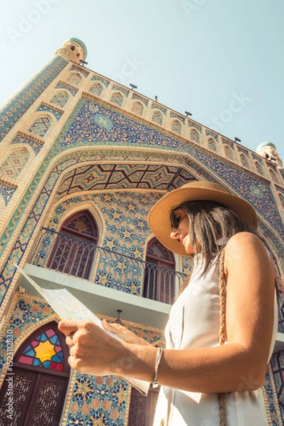 Fototapeta Smiling female tourist with map by colorful islamic architecture Persian-style mosque building. Travel sightseeing planning, cultural discovery, solo journey, heritage adventure