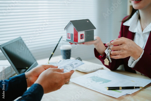 Obraz Asian female real estate agent in red shirt introduces house offer. Confident and friendly real estate agent presents small model home to large client.