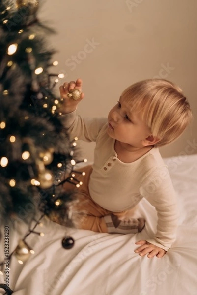 Fototapeta Blond toddler in warm home clothes curiously explores Christmas tree decorations reaching for golden ornament with fascination and wonder in cozy festive atmosphere Little child reaches for golden bal