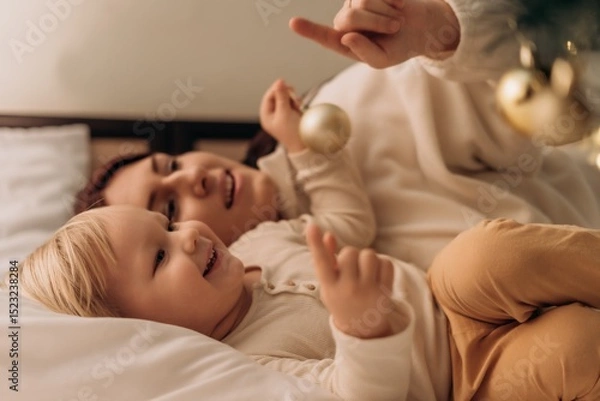 Fototapeta Smiling toddler lies in bed beside mother pointing at Christmas tree lights while holding ornament sharing magical moment of love togetherness warmth joy and connection in modern motherhood
