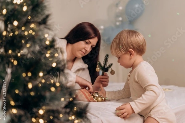 Fototapeta Mother and toddler enjoy meaningful celebration while decorating with baubles and fir sprigs near Christmas tree sharing quiet joy in warm holiday atmosphere full of love and seasonal connection