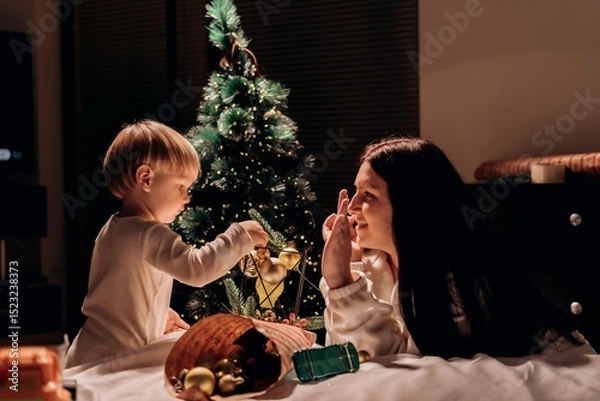 Fototapeta Smiling modern mother plays with son while decorating festive tree with golden ornaments sharing warmth and playful joy in a simple evening moment of holiday connection and gentle parenting