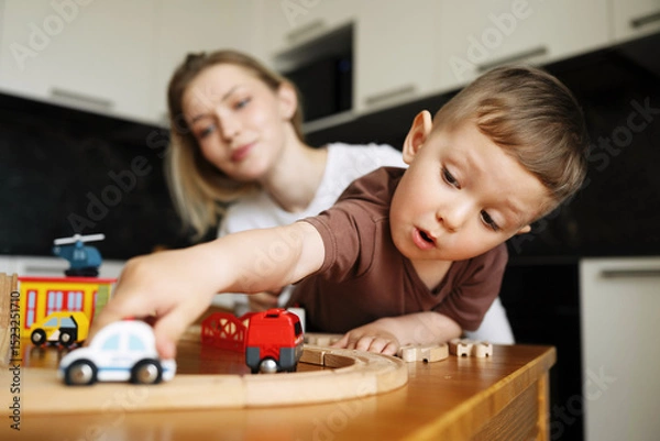 Fototapeta Little boy playing with excitement while pushing a toy car on a wooden train track, with his mother watching in the background at home.