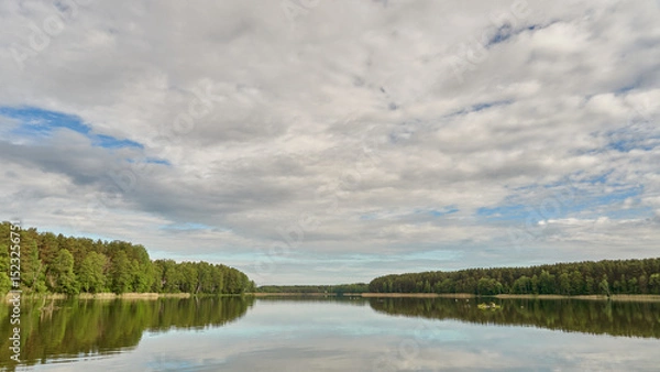 Fototapeta Sunny summer landscape on the lake. Reflection of the sky and clouds in the water. A valley of green trees on the shore of a lake. A concept for travel and camping.
