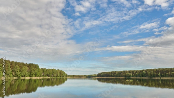 Fototapeta Sunny summer landscape on the lake. Reflection of the sky and clouds in the water. A valley of green trees on the shore of a lake. A concept for travel and camping.