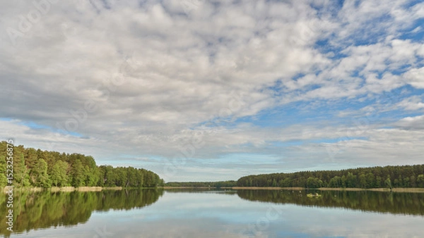Fototapeta Sunny summer landscape on the lake. Reflection of the sky and clouds in the water. A valley of green trees on the shore of a lake. A concept for travel and camping.