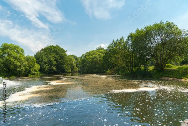 Fototapeta Sunny summer landscape on the lake. Reflection of the sky and clouds in the water. A valley of green trees on the shore of a lake. A concept for travel and camping.