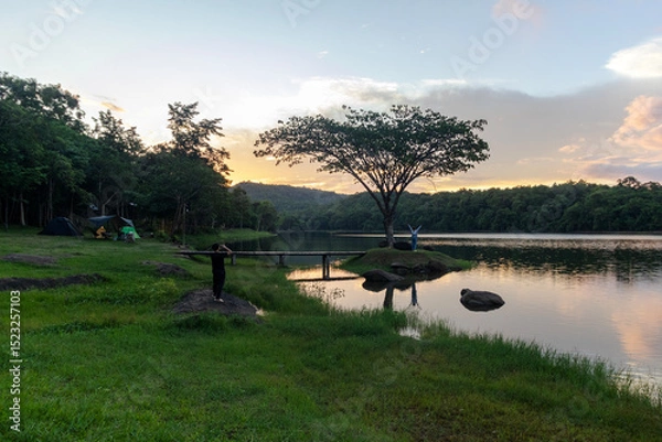 Fototapeta Tourists enjoy photographing the sunset at Pha Daen Forest Park, a tiny tree-covered island viewpoint with a beautiful bridge and warm golden hour light.
