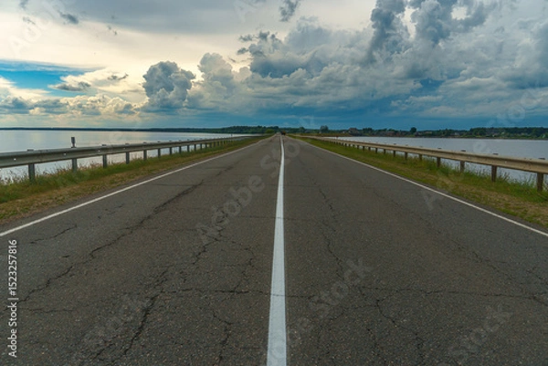 Fototapeta An empty asphalt road runs through the river. Thick gray clouds over the highway along the lake.