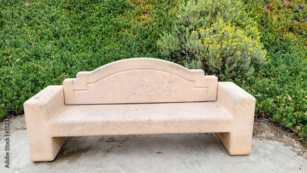 Fototapeta Beige colored concrete park bench with a curved backrest set against a backdrop of green Ice Plant and Bladderpod near cliff slope, San Diego, California