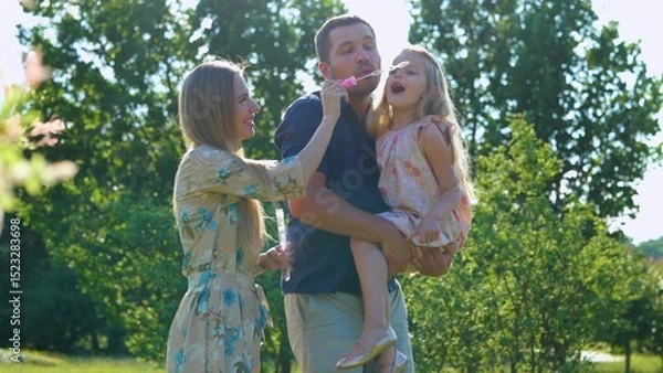 Fototapeta Joyful family spending summer day together outdoor playing with soap bubbles. Parents play with child on a walk. Girl blowing bubbles with father and mother in park. Childhood concept