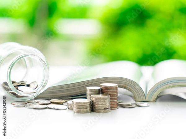 Fototapeta An open book and a jar of coins sit next to a small pile of loose change.