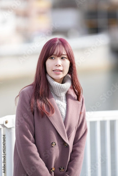Fototapeta On a chilly December morning, a Japanese woman in her early twenties with long pink hair and a pink trench coat walks a quiet riverside path in a well-kept city, enjoying the gentle winter charm.