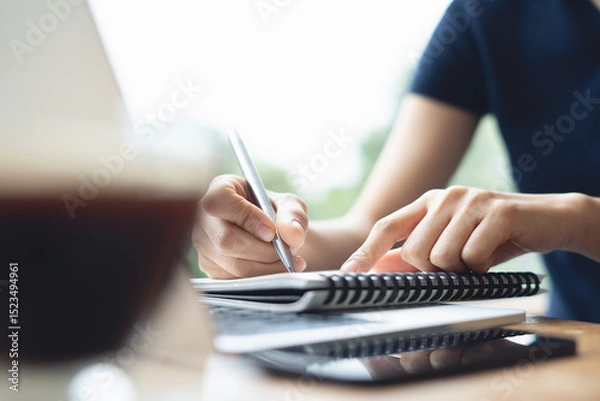Obraz Business woman sitting at table in cafe, writing in notebook with laptop computer, mobile phone on table. Freelancer working in coffee shop. Student learning online, doing research and taking note