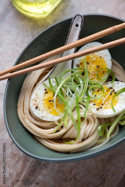 Fototapeta Udon noodles with boiled chicken egg and spring onion in a green bowl, vertical shot on a brown granite surface, middle close-up