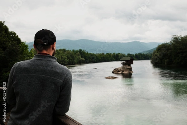 Obraz Man in a grey fleece jacket looking at the iconic house on the Drina River from a wooden viewpoint. Rear view. A popular tourist destination. Serbia country in spring season.