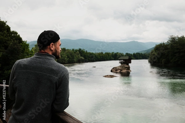Obraz Man in a grey fleece jacket looking at the iconic house on the Drina River from a wooden viewpoint. Rear view. A popular tourist destination. Serbia country in spring season.