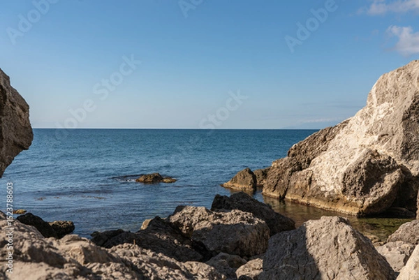 Fototapeta Coastal scene with clear blue water gently lapping against rugged rocks. Rocky coast of Black Sea. Cape Alchak. Sun casts warm glow, highlighting textures of stones and tranquil sea. Sudak, Crimea