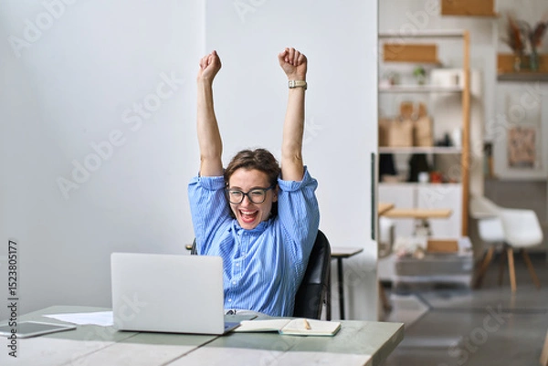 Obraz Excited office worker winner using laptop computer celebrating goal achievement. Happy young woman student or employee winning online getting good news in email raising hands feeling euphoric.