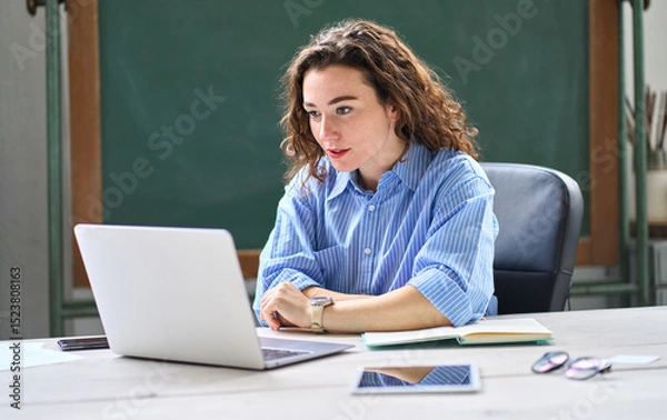 Obraz Female online teacher looking at laptop talking on virtual distance class in classroom or webinar web training working in office. Young business woman leading hybrid conference online video call.