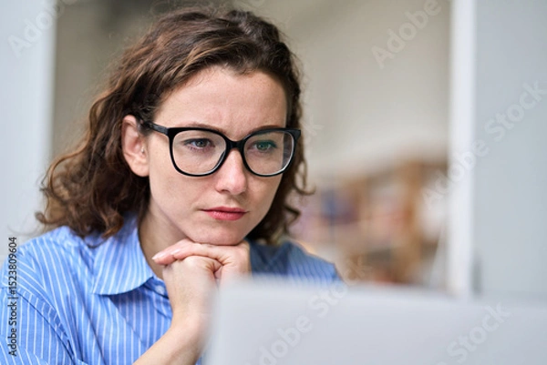 Obraz Busy young serious professional business woman employee or student wearing glasses using laptop watching online webinar or training web course, looking at computer, thinking, elearning. Close up
