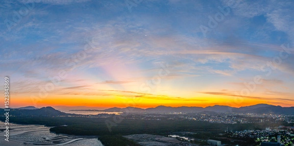 Obraz A stunning panoramic view of a coastal city at sunset at Phuket city, showcasing vibrant hues of orange and blue in the sky, with mountains, and urban lights creating a harmonious blend of natural